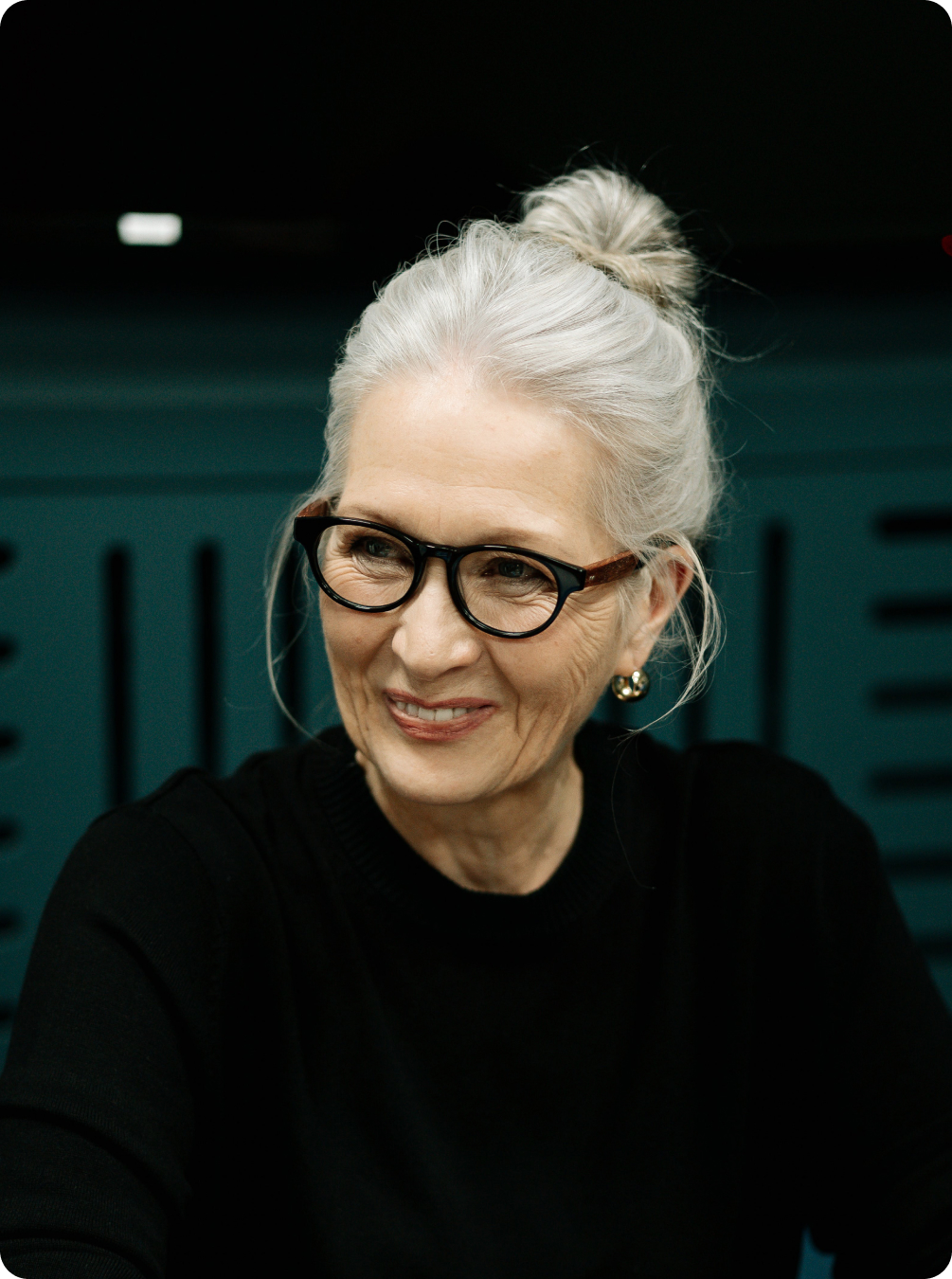 A person with gray hair tied up in a top knot wearing a black top in an office.