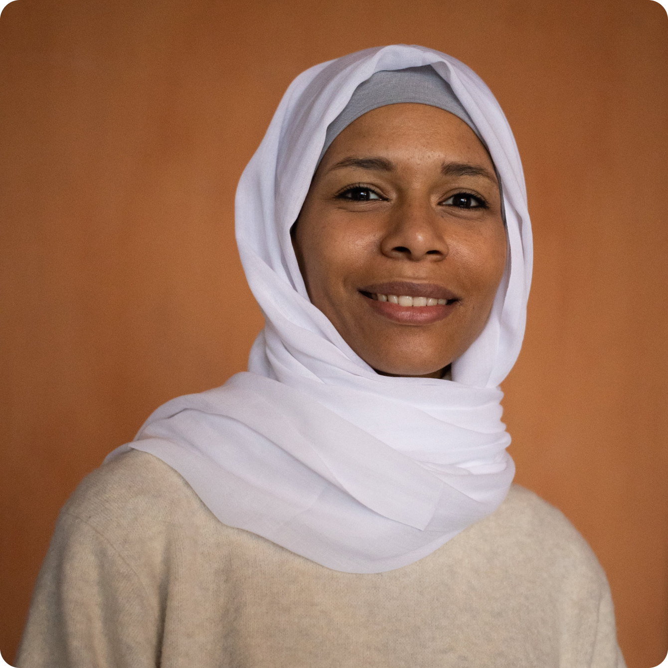 A person wearing a white hijab and a beige top against an orange background.